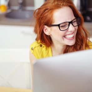Head and Shoulders of Young Woman with Red Hair Wearing Eyeglasses and Laughing Joyfully While Working on Computer with Over-Sized Monitor at Home in Kitchen