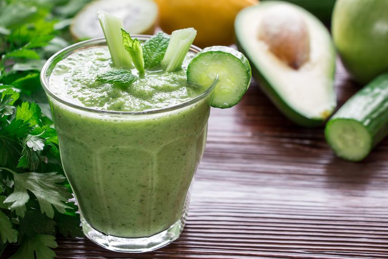 Left glass of green vegetable smoothie near ingredients celery, avocado, cucumber, apple, kiwi and herbs, right empty space on wooden background. Green vegetable smoothie and ingredients. Horizontal.