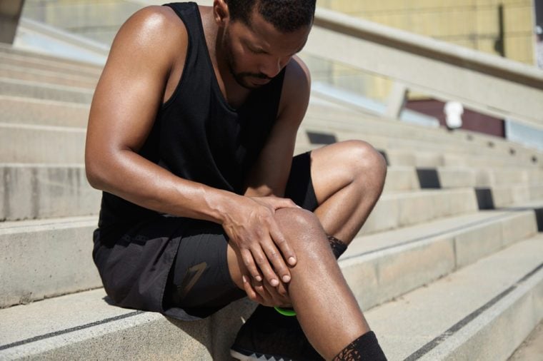 Handsome muscular male jogger wearing black training outfit touching his knee in pain with clasped hands, having sprain or rupture in his muscles after exercising outdoors. Sports injury concept