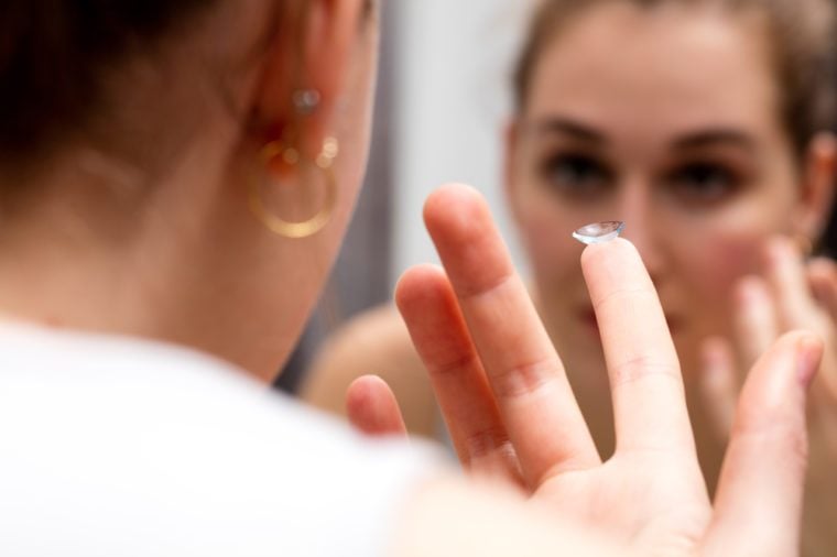 closeup of young woman looking at herself in the mirror to change her contact lens, holding one corrective eye care product, female face in a blurred background