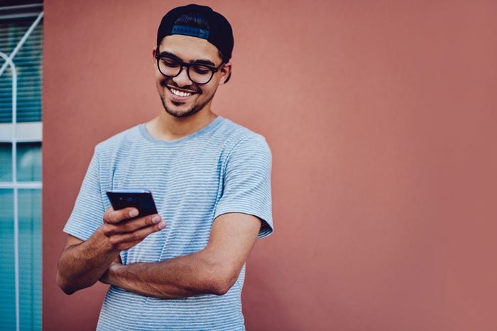 Cheerful young man in trendy baseball cap watching funny video on mobile phone using 4G connection, smiling hipster guy reading good news from networks standing on wall promotional background