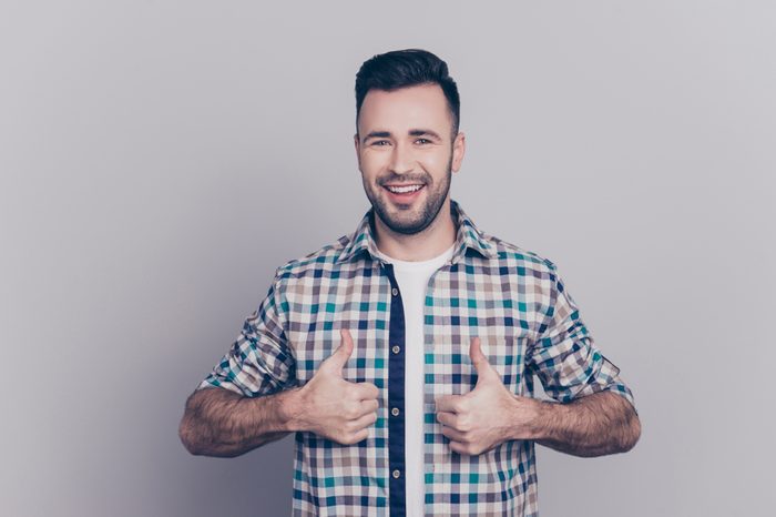 Portrait of smiling, bearded, perfect man showing two thumbs up in checkered shirt over grey background
