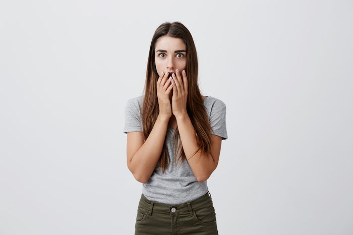 Body language. Portrait of young attractive charming caucasian girl with dark long hair in casual gray t-shirt and jeans clothing mouth with hands, looking in camera with raised eyebrows and shocked