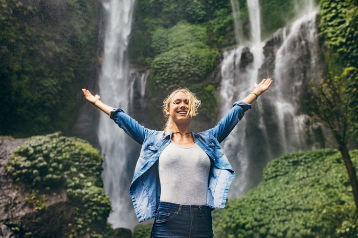 Happy young woman spreading hands enjoying nature with waterfall in background. Caucasian female standing in front of a waterfall with her arms outstretched.