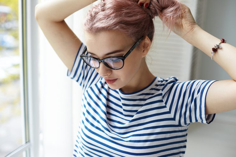 Morning routine. Beautiful young female with pinkish hair wearing blue and white striped top and trendy eyeglasses standing at window and doing hairstyle, going out to have coffee with friends