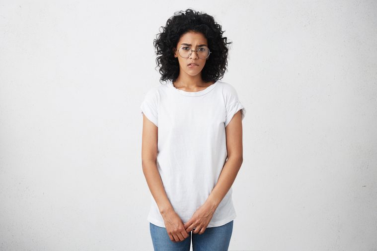 Shy and timid African American student girl wearing glasses, white t-shirt and jeans having indecisive confused look while answering to teacher's question in class, biting lips, feeling nervous