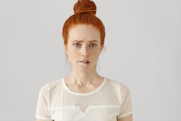 Confused or puzzled beautiful young Caucasian woman with ginger hair frowning, biting her lower lip after having done something wrong, looking at camera with guilty and apologetic facial expression