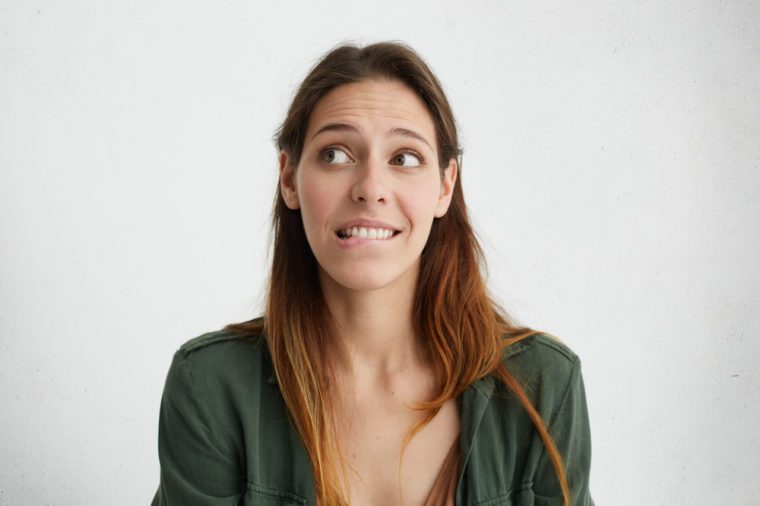 Indoor portrait of shy pleasant-looking female looking embarrassed aside biting her lower lip wanting to say something, but not having courage to do it. Woman feeling her guilt looking confused