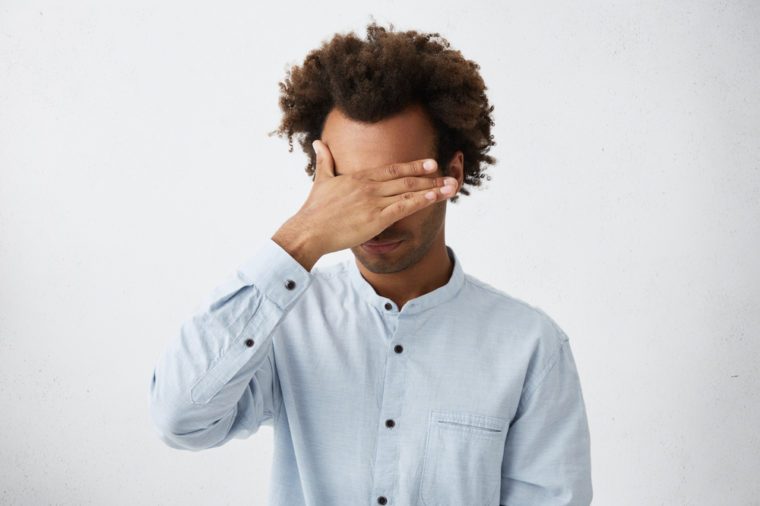 Unrecognizable unshaven African American man with tousled hair making facepalm gesture, covering eyes while feeling ashamed, standing against blank studio wall background with copy space for content