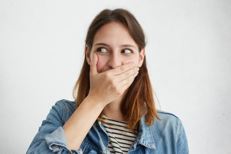 Pretty female covering her mouth while giggling looking aside posing against white studio background. Stylish woman dressed in jean jacket trying to control her emotions, bursting into laughing