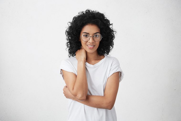 Beautiful young dark-skinned female wearing stylish round glasses and black casual t-shirt, making awkward gesture, touching her neck during intense conversation with someone, trying to smile