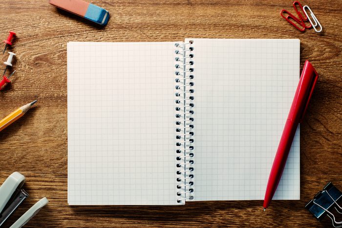 Red pen ready for writing on an open spiral notebook with blank pages with math pattern surrounded by school supplies on a wooden table, high-angle close-up