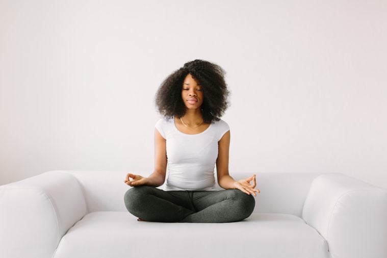 An African American young women sitting in the lotus position on white bed.