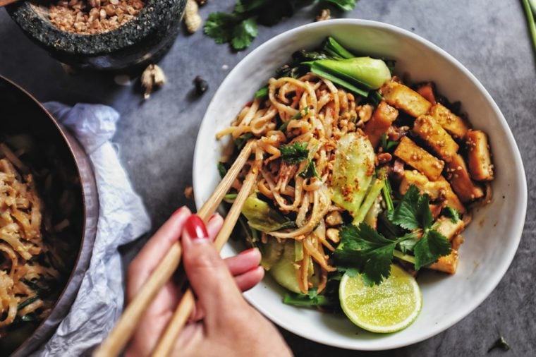     Udon with Padthai sauce, Healthy Vegetarian/Vegan menu;  Padthai noodles with smoked tofu and mixed vegetables - chinese baby bok choy, garlic chives, shallot and crushed peanut topping.  holding chopstick.