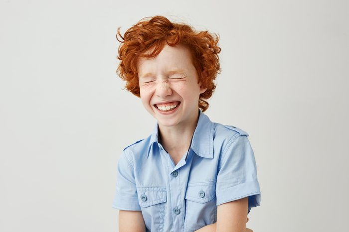 Close up portrait of cheerful little kid with curly ginger hair and freckles laughing with closed eyes after hearing funny joke from friend.