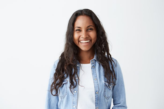 Beautiful dark-skinned female with long brunette hair and broad happy smile wearing denim shirt enjoying good positive news concerning her promotion at work, posing isolated against white blank wall