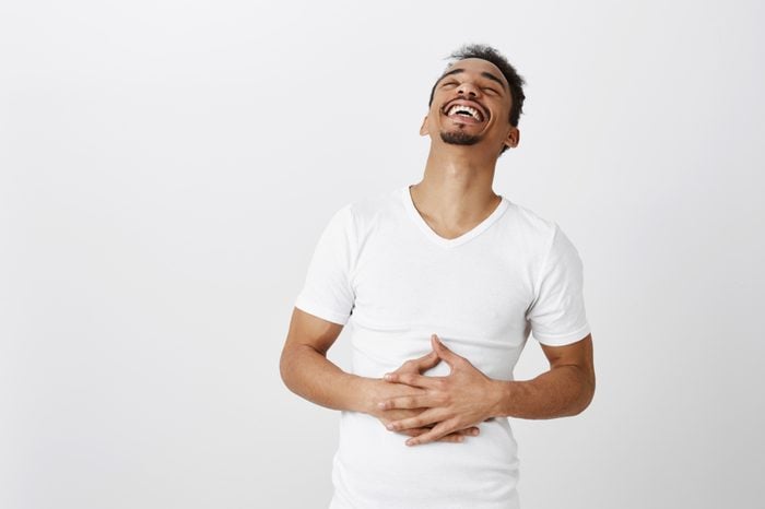 What nice time to live. Portrait of carefree attractive male student in white t-shirt, hearing funny joke, laughing out loud, feeling pain in belly muscles from chuckle, standing over gray wall