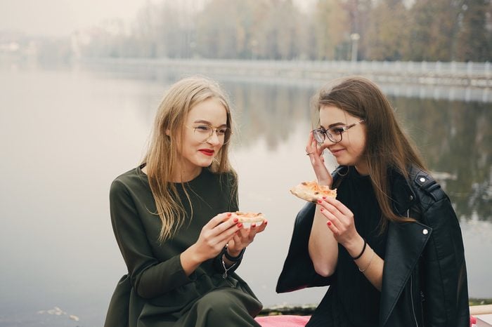 две молодые и стильные девушки гуляют в осеннем парке у воды и едят пиццу