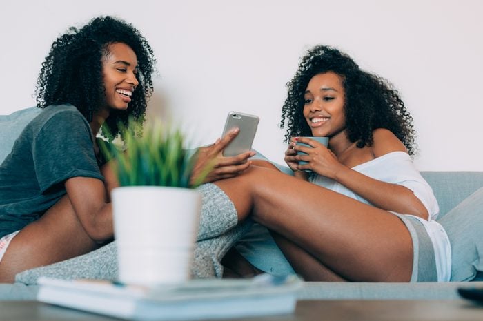 Счастливые молодые две чернокожие женщины сидят на диване, смотрят в мобильный телефон и пьют кофе