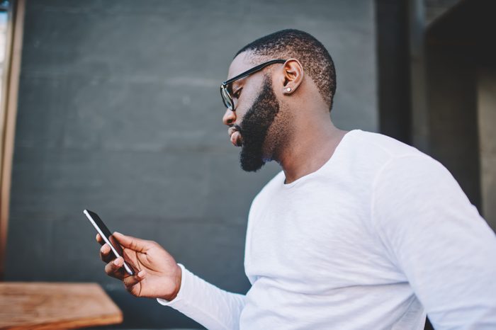 Side view of handsome african american guy reading message on phone while standing outdoors in, dark-skinned hipster in shirt with copy space for brand name or label installing application on mobile