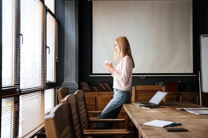 Image of pretty young lady standing in cafe drinking coffee and looking aside while work with laptop.