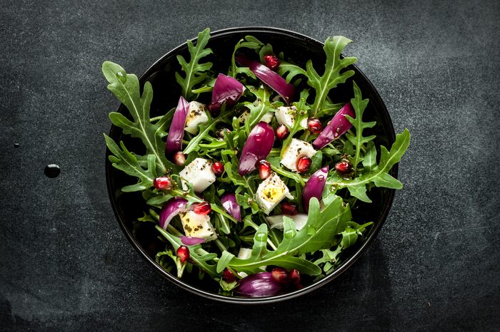 Fresh spring salad with rucola, feta cheese, red onion and pomegranate seeds in black bowl on chalkboard background. 