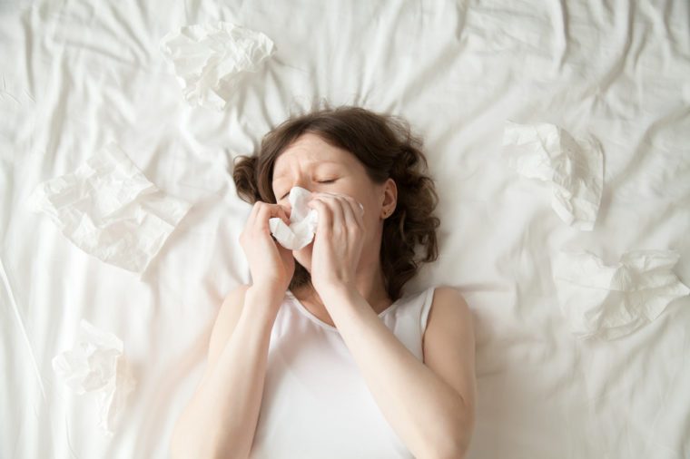 Young depressed or sick with flu model lying on the bed with closed eyes and blowing her nose into tissue. View from above