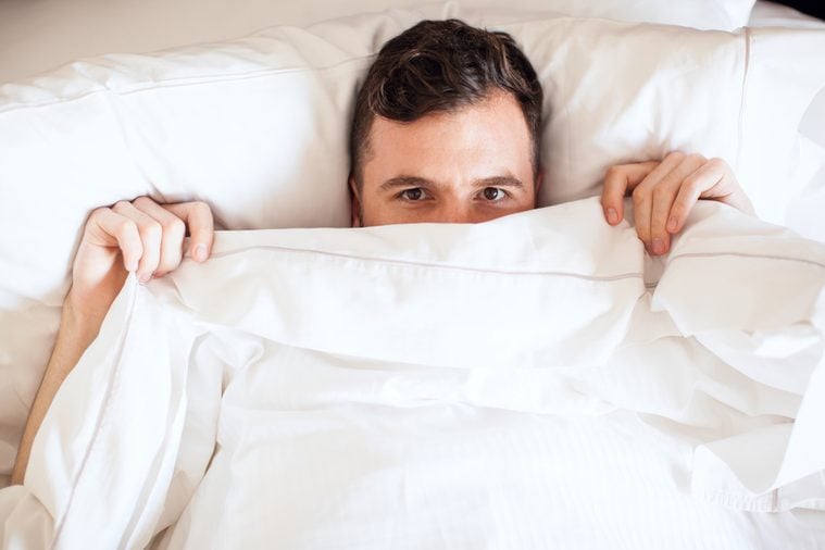 Portrait of a young man pulling the sheets of his bed up and peeking over them