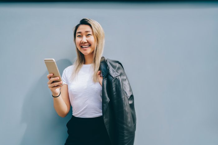 Cheerful blonde asian female holding mobile phone and posing on copy space background, happy Chinese woman dressed in mock up white t-shirt sending messages on smartphone using internet connection