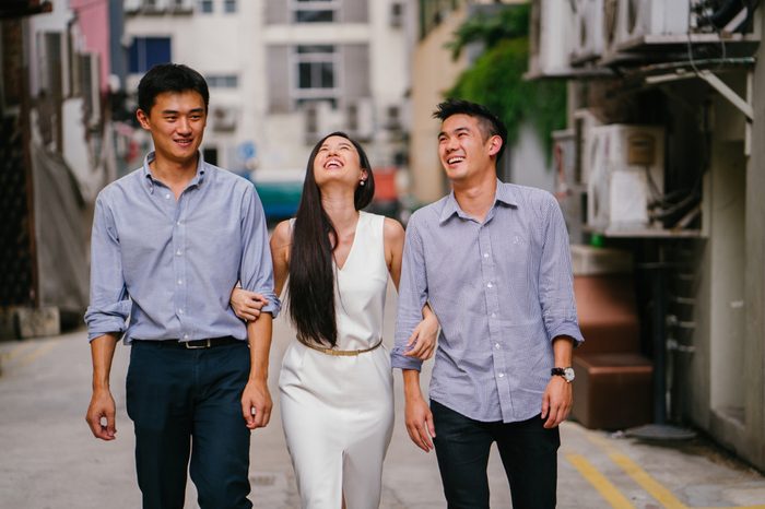 A young, attractive Chinese woman walks along a street in Asia during the day with her arms crossed with two young Chinese men. They are all laughing as they walk. 