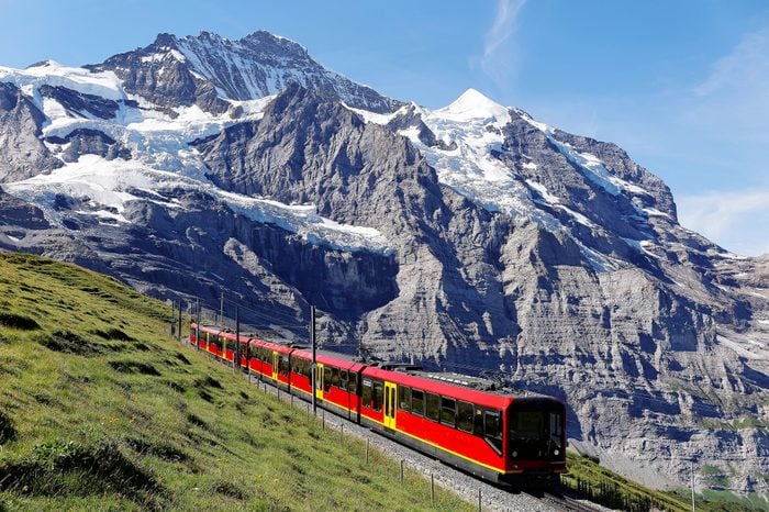 A sightseeing train travels on the famous Jungfrau Railway from Jungfraujoch (Top of Europe) to Kleine Scheidegg by a green grassy hillside on a sunny summer day, in Bernese Oberland, Switzerland