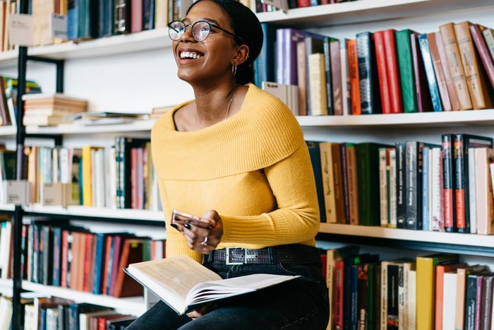Positive african american young woman in stylish eyeglasses for vision correction laughing while received funny notification on modern smartphone device sitting with literature book in modern library