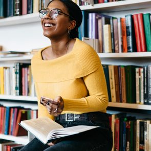 Positive african american young woman in stylish eyeglasses for vision correction laughing while received funny notification on modern smartphone device sitting with literature book in modern library