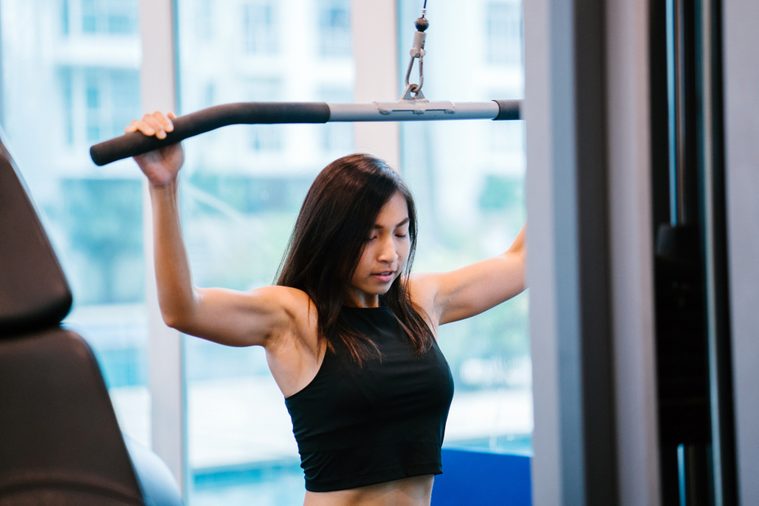 Portrait of a young Chinese Asian woman doing strength training at the gym. She is tanned, toned, muscular and fit and is focused on lifting the weights. 