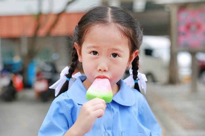 Adorable little Asian child girl in school uniform sucking or eating ice-cream in the park
