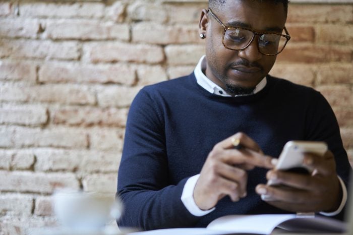 Cropped image of afro american international student in spectacles browsing internet websites on smartphone in searching useful languages seminars for studying remote using free wifi in coffee shop