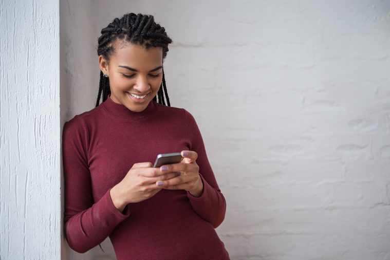 Smiling Black Woman Texting on Smartphone by Wall