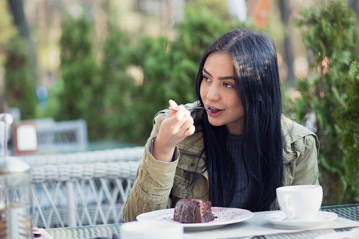 Close up portrait of a young woman eating dessert chocolate ice cream licking spoon outdoors in summer terrace coffee shop. Side view