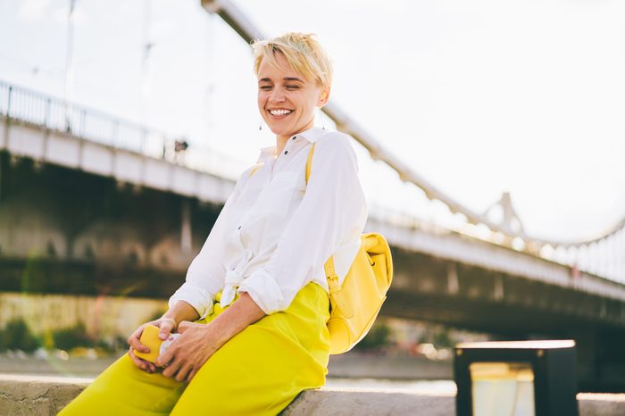 Happy gorgeous female dressed in stylish yellow casual clothing laughing sitting in urban setting near bridge.Positive young woman with trendy backpack having fun outdoors in city centre