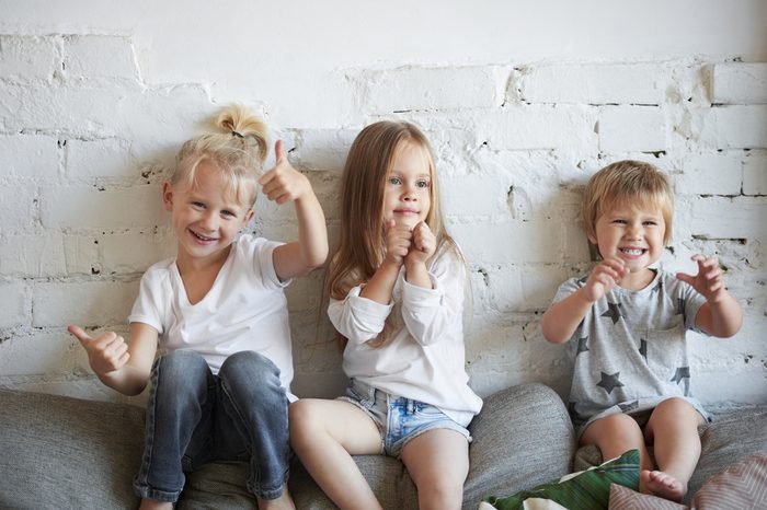 Candid shot of three siblings posing in living room, sitting on top of sofa at white brick wall: blond boy showing thumbs up gesture, girl in the middle pumping fists and their little sister grimacing