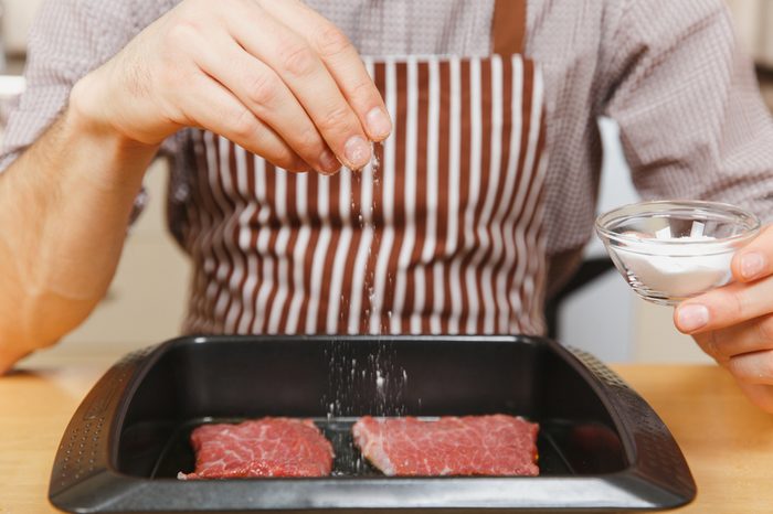 Close up man in apron sitting at table with vegetables, sprinkling meat seasoning in black baking tray, cooking at home preparing stake from pork, beef or lamb, in light kitchen with wooden surface