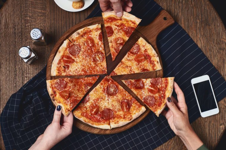 Flatlay. Close-up of people hands taking slices of pepperoni pizza from wooden board. Table served with black textile napkin. Smartphone on table. People eat fast food in cafe.