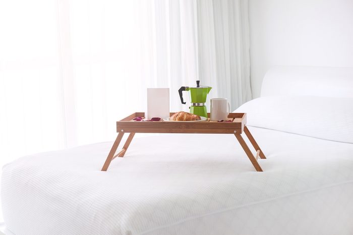 Breakfast wooden tray with coffee percolator, white blank card and croissant on bed. Light from window inside the room