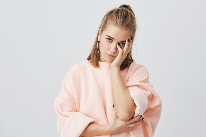 Bored European girl student wearing stylish pink sweatshirt touching face with hand, looking annoyed, tired of listening uninteresting stories. Body language