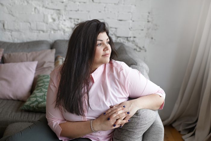 Indoor shot of beautiful plus size fashion female model with straight black hair relaxing on comfortable gray sofa by the window at home, having pensive thoughtful expression, wearing pink shirt.