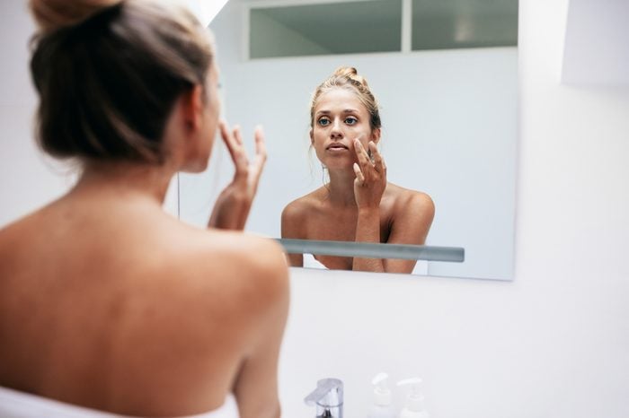 Woman in bathroom looking in to mirror and applying cream on her face . Beautiful female pampering her facial skin.