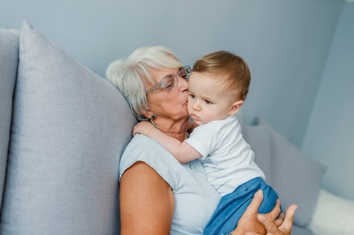 Grandmother is kissing her baby grandson - indoors scene. Proud Grandmother Holding Baby Grandson. Senior woman hold little baby boy. Multi generation family playtime