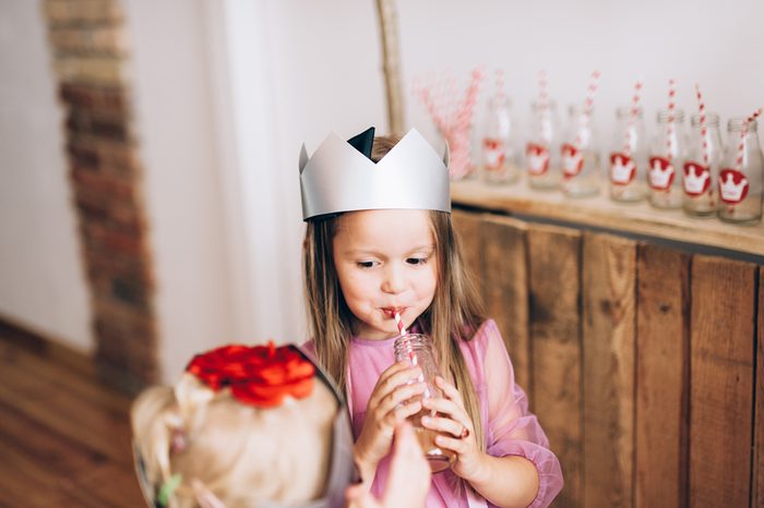 Young little girl wearing princess crown and pink dress, smiling, drinking juice and celebrating her birthday