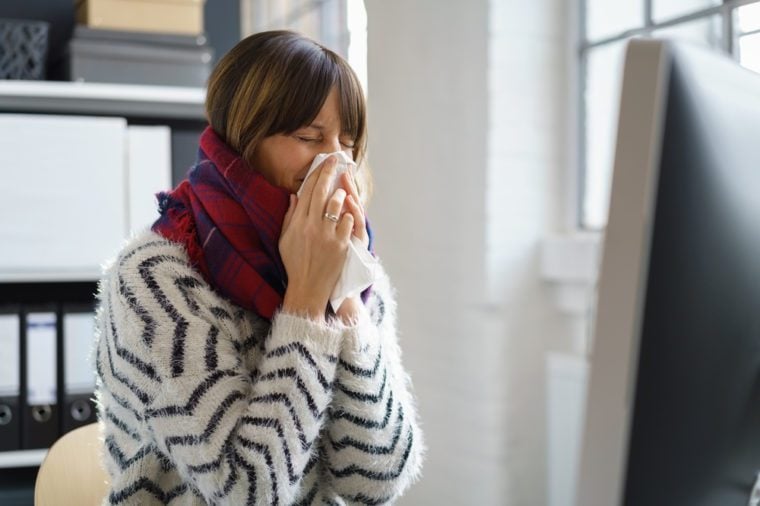 Sick businesswoman with seasonal winter flu blowing her nose in a tissue as she sits at her desk in a warm thick woollen scarf