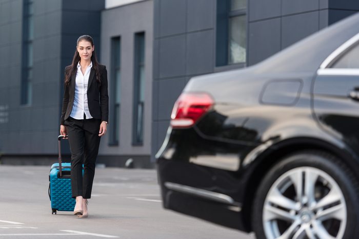 Young beautiful woman in pant suit walking towards passenger car with suitcase